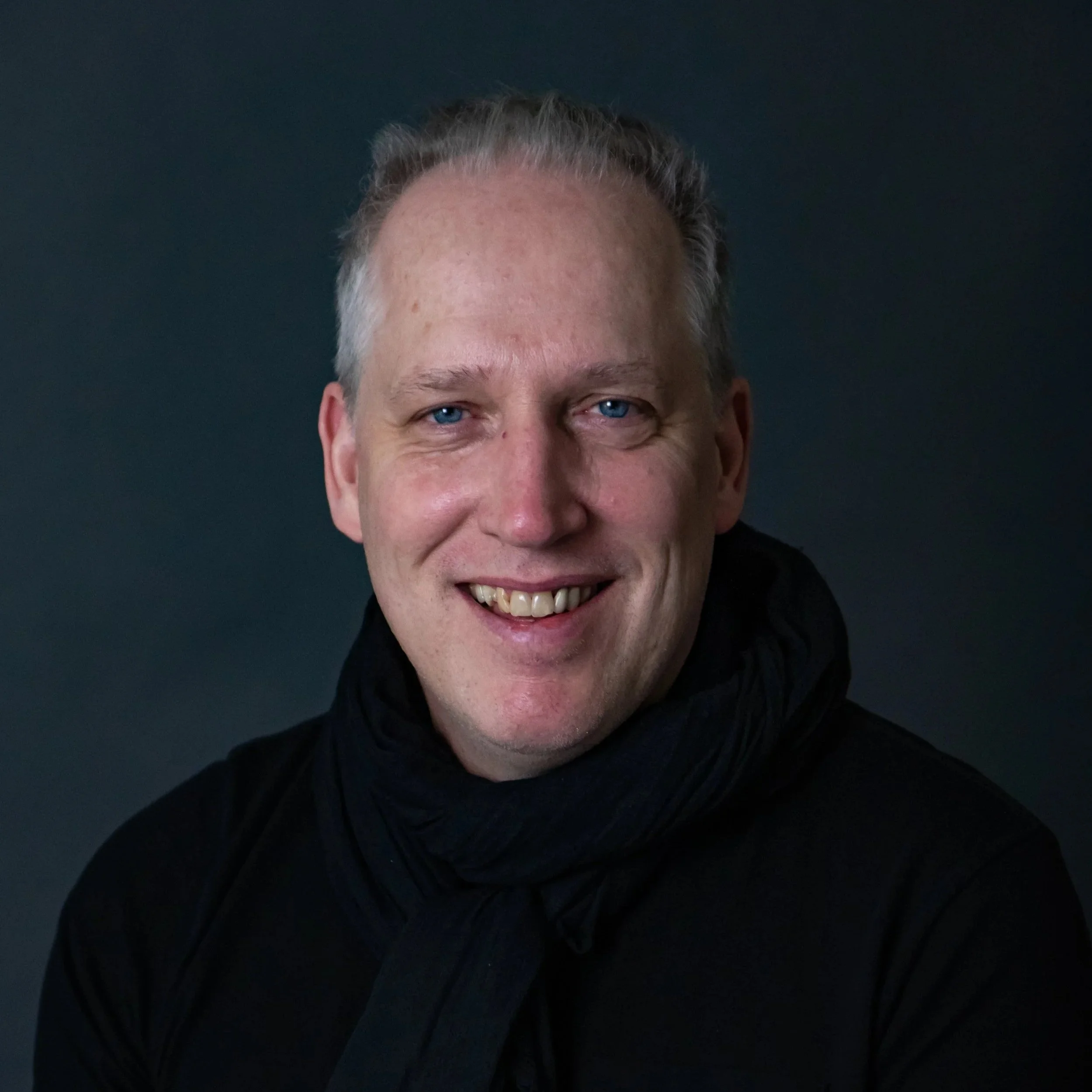 A smiling man with light blond hair, blue eyes, and fair skin, wearing a black top and a black scarf, against a dark background.