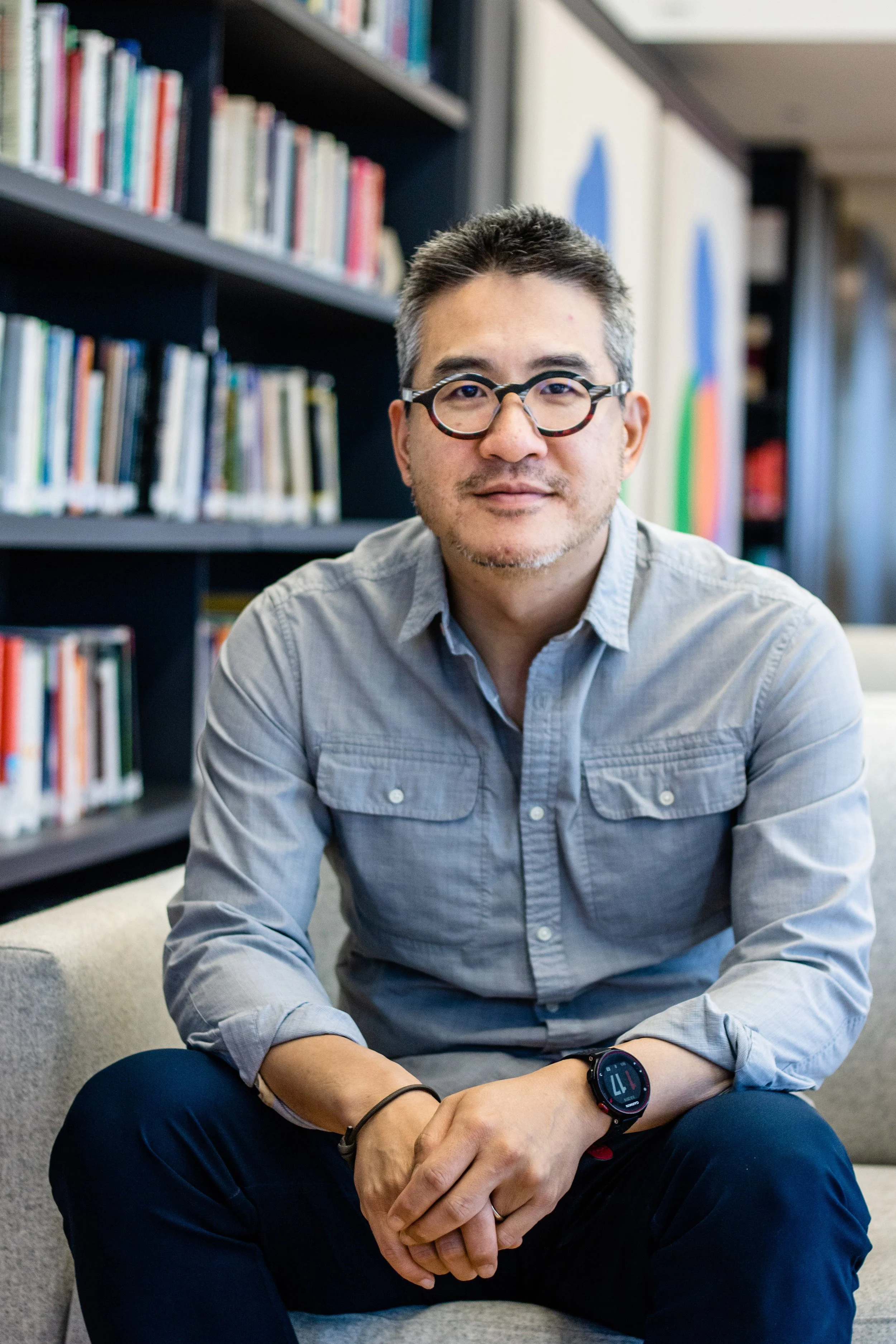 A man wearing glasses, a gray button-up shirt, and a smartwatch sitting on a beige couch in a library with bookshelves filled with books behind him.