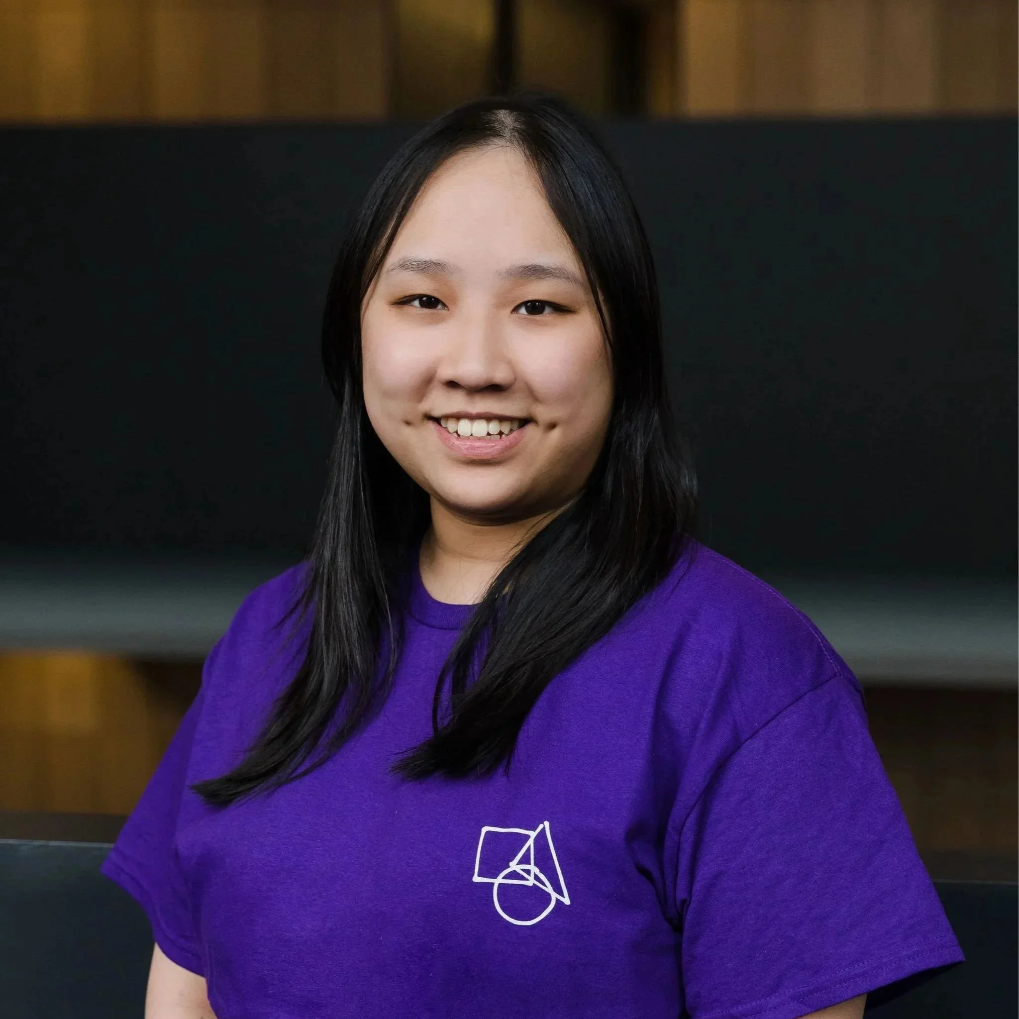 A young woman with black hair smiling, wearing a purple T-shirt with a white geometric logo, standing indoors in front of a dark background with wooden accents.