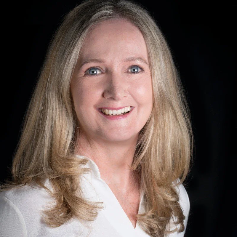 A woman with blonde hair, blue eyes, and fair skin, smiling against a black background, wearing a white top.