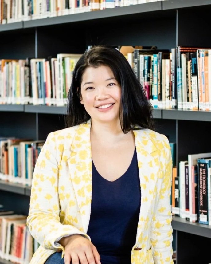 A woman with black hair, wearing a yellow floral blazer and navy blue top, sitting in front of bookshelves filled with books, smiling at the camera.