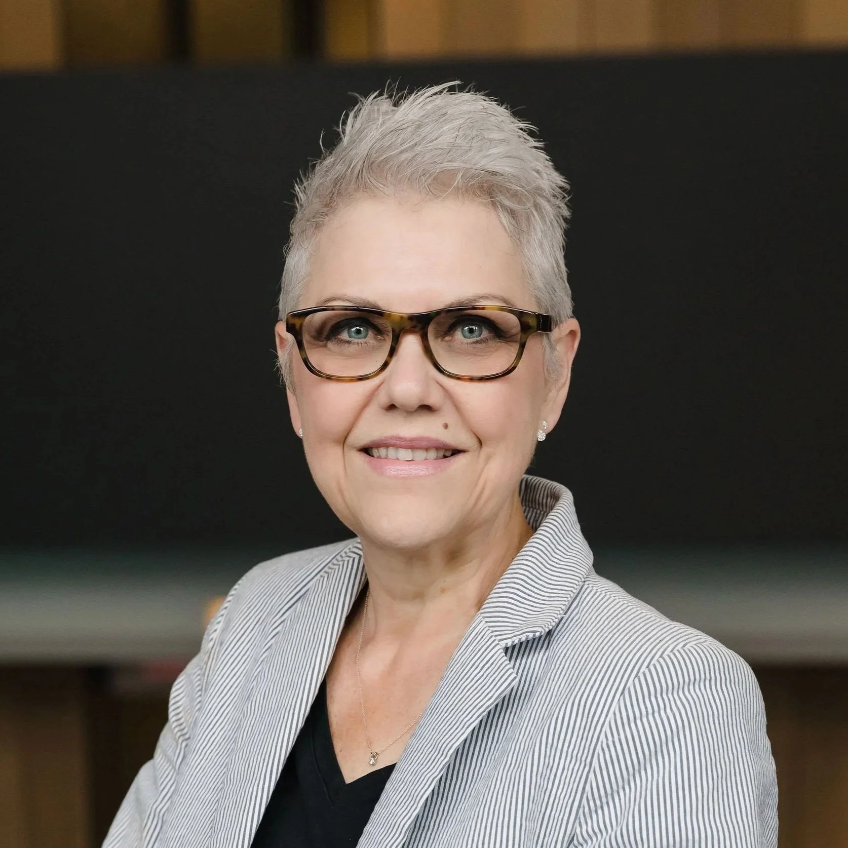 A woman with short gray hair, wearing glasses, a striped blazer, and a black top, smiling in front of a black background.