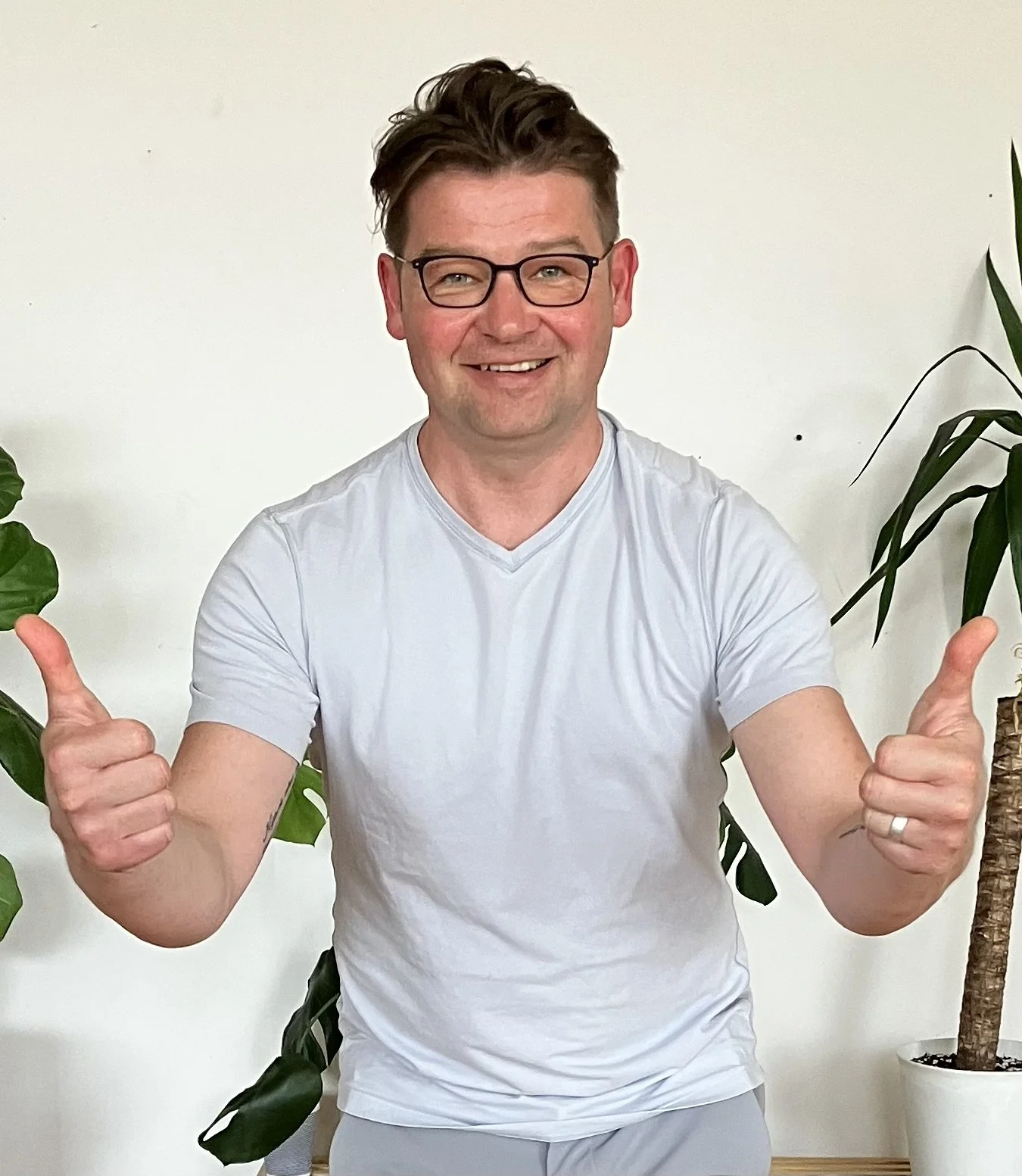 Man with short dark hair and glasses smiling and giving two thumbs up, standing in front of a white wall with green indoor plants on either side.