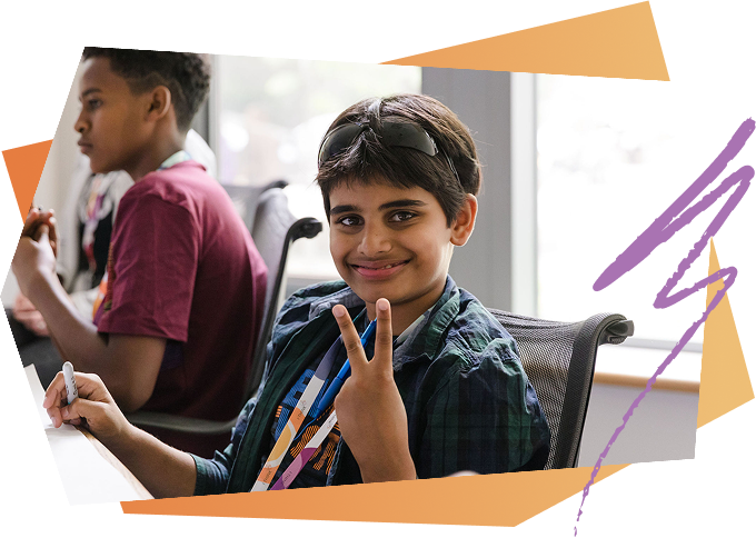 A young boy smiling and making a peace sign while sitting at a desk in a classroom, with another student in the background.
