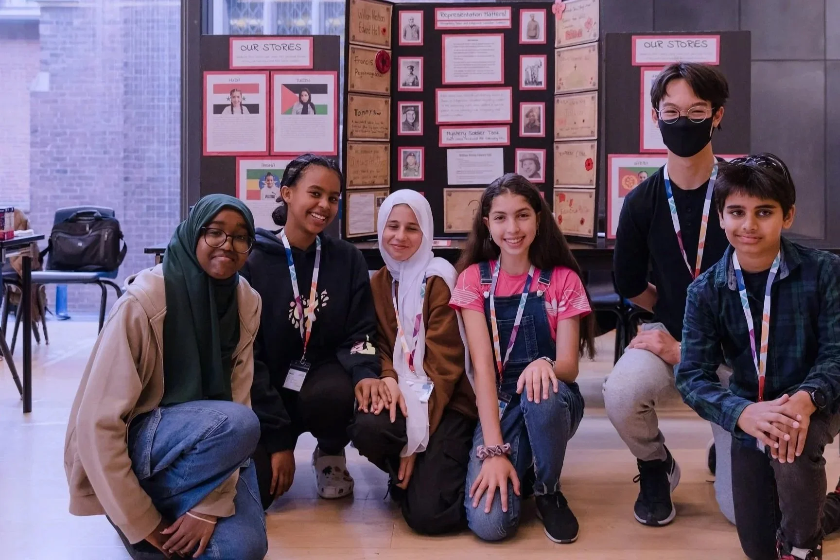 Group of six students smiling indoors, kneeling and standing in front of a display board with school projects and posters.
