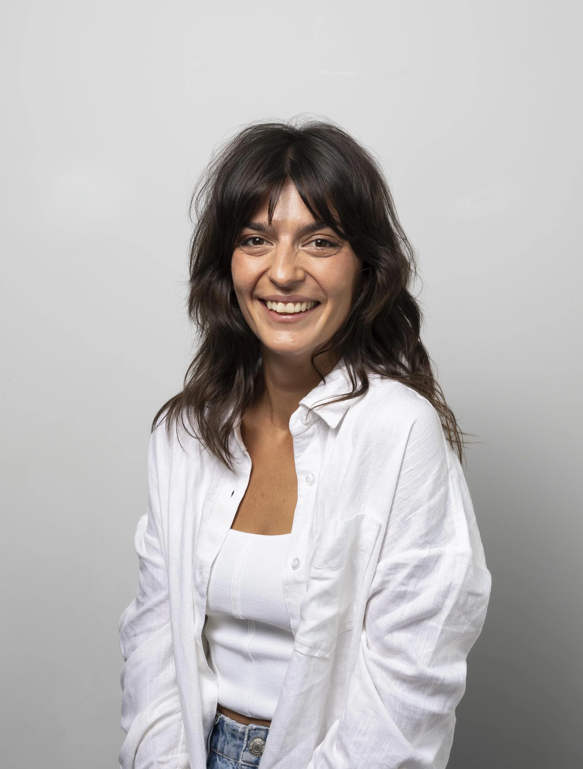 A woman with shoulder-length wavy dark hair, smiling, wearing a white button-up shirt over a white tank top, standing against a plain light gray background.