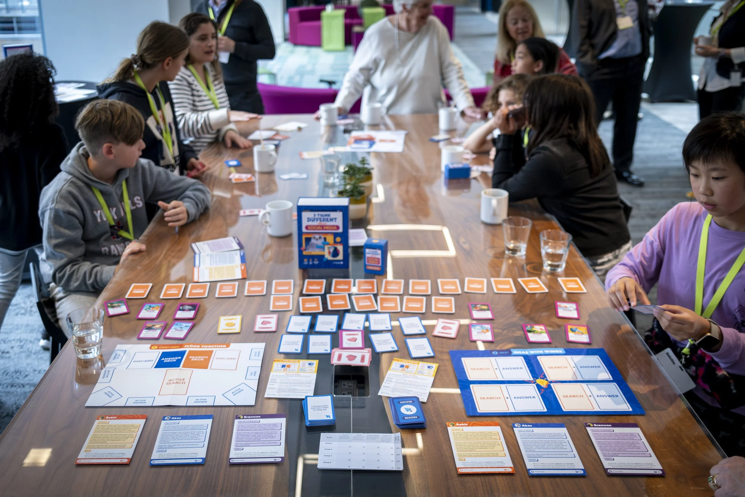 Children and adults gathered around a large wooden table playing a tabletop game involving cards, tiles, and game boards, in a modern indoor setting.