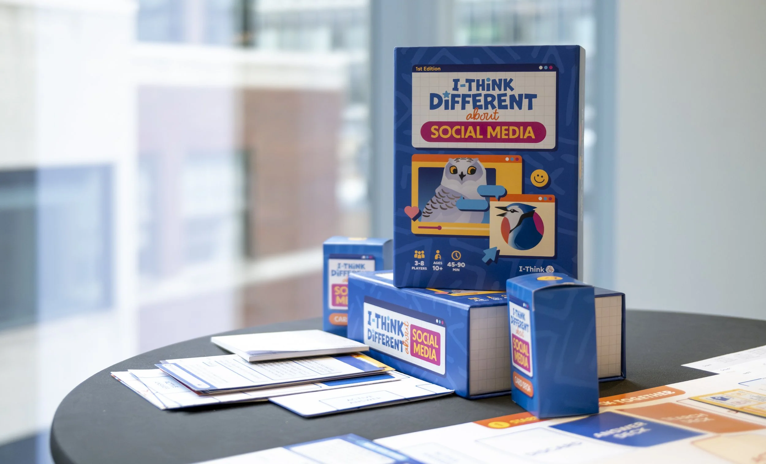Boxes of the game 'I-Think Different about Social Media' on a table with game materials and instructions, with a blurred background.