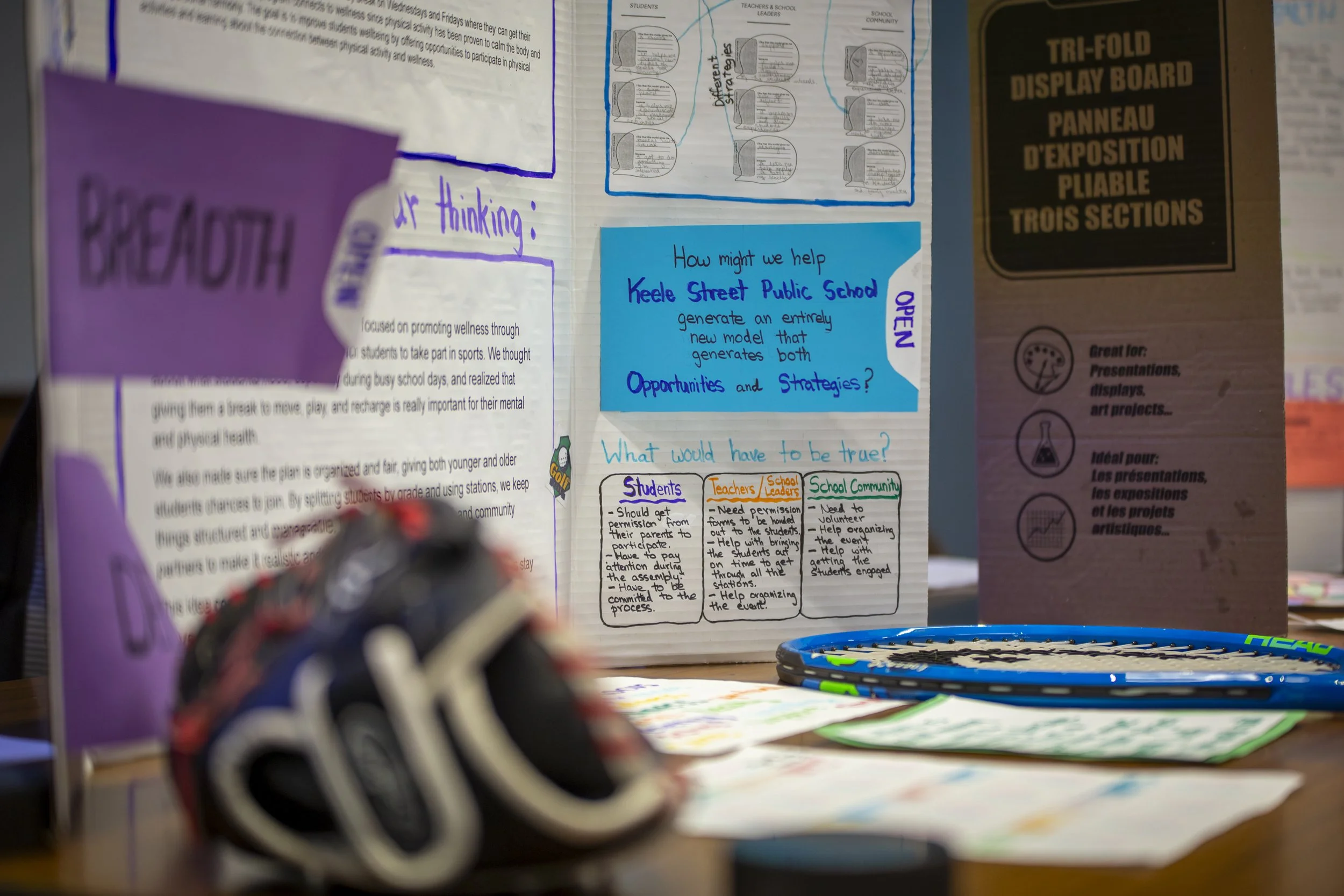 An educational display board and materials on a table, including a blue poster with questions about helping Keele Street Public School, a tri-fold display board, and tennis rackets, with handwritten notes and printed information about community engagement and student and teacher permissions.