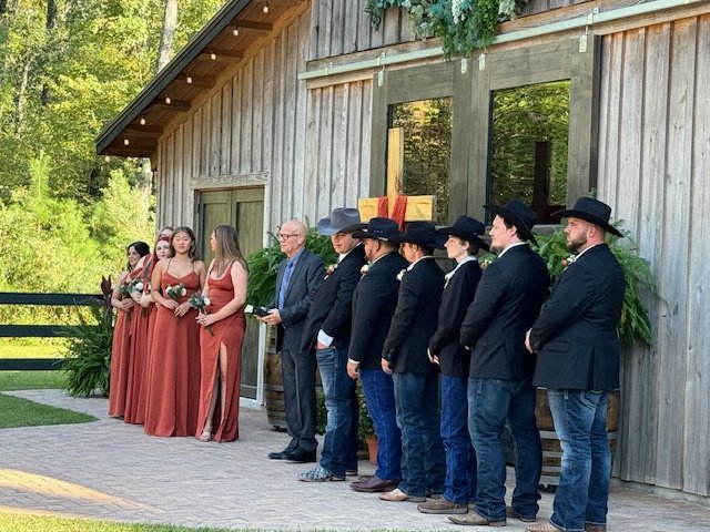 A wedding ceremony outdoors with bridesmaids in burgundy dresses and groomsmen in cowboy hats and suits standing in front of a rustic wooden building.