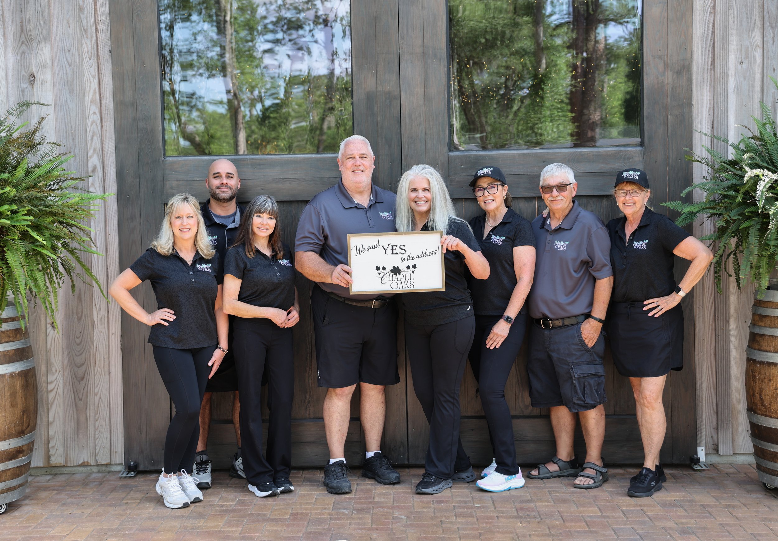 A group of nine smiling people posing in front of a wooden barn door with trees reflected in the windows behind them, some wearing black shirts and hats with a logo, one person holding a sign that says 'We said YES to the address! CHAPEL OAKS', and two potted ferns flanking the group.