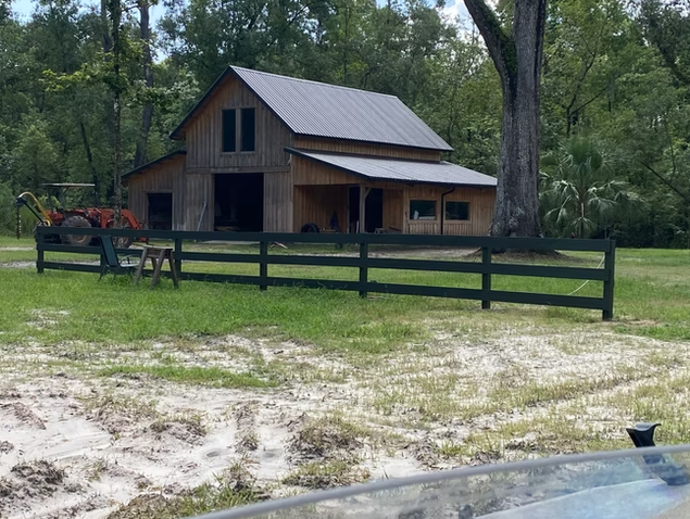 Wooden barn with a metal roof, surrounded by trees, with a black fence and some equipment in front.