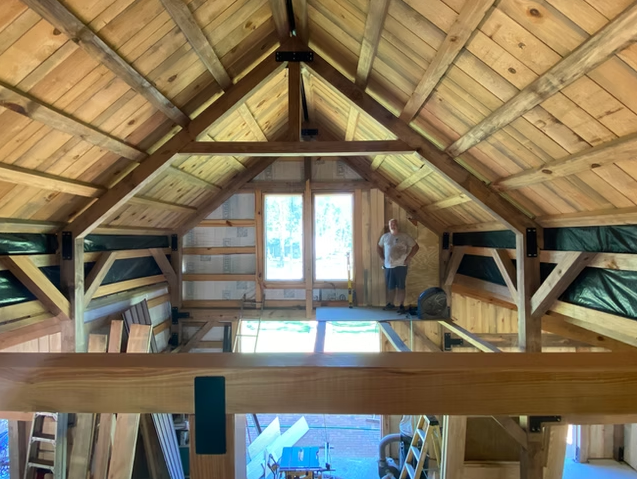 A man inside a wooden attic or loft under construction, with exposed wooden beams and framing, and a large window at the far end allowing natural light.