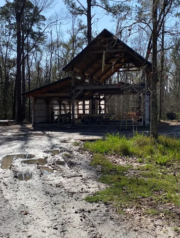 Old, tilted wooden barn structure in a wooded area with a muddy path in the foreground.