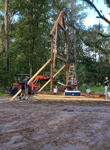 Construction workers building a wooden structure outdoors with a tractor nearby and construction tools around, surrounded by trees.