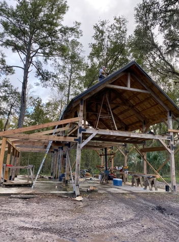Construction site of a wooden building with workers on the roof, scaffolding supports, surrounded by trees.