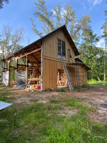 A two-story wooden house under construction in a wooded area, with scaffolding and construction materials around it.