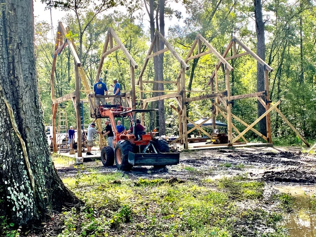People building a wooden playground structure in a wooded outdoor area.