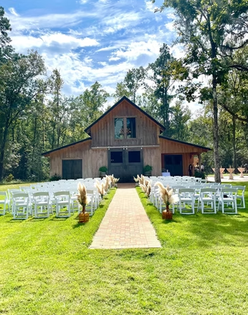 Outdoor wedding setup with rows of white chairs on each side of a brick pathway leading to a rustic barn decorated with plants and pampas grass, surrounded by tall trees under a partly cloudy sky.