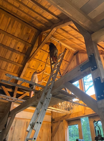 Two people working on a wooden ceiling in a rustic room, using ladders and tools, with large windows and a chandelier visible.