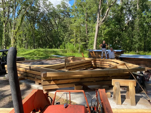 Two people working on a boat construction site outdoors, with a partially built wooden boat frame and various tools, surrounded by trees and greenery.