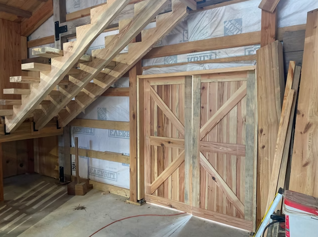 Unfinished interior of a wooden building with a staircase, a barn-style door, and construction materials.