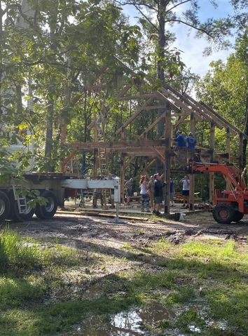 Construction workers building a wooden house with a crane and truck on a wooded lot.