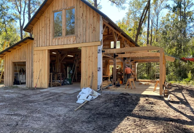A two-story wooden house under construction, with construction tools and materials outside, surrounded by trees.