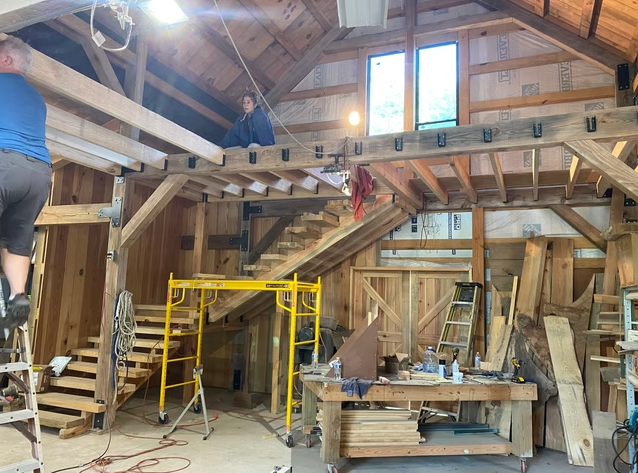 Interior of a wooden house under construction with unfinished walls and ceiling, scaffolding, a work table with tools, and two people working on the structure.