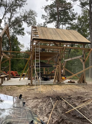 Wooden house under construction in a wooded area with scaffolding, ladder, and construction tools.