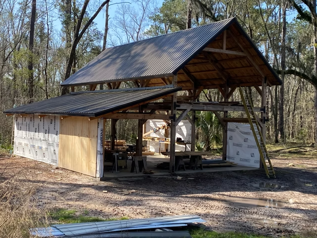 A partially built outdoor shelter under construction in a wooded area, with a metal roof and wooden framework, surrounded by construction materials and tools.