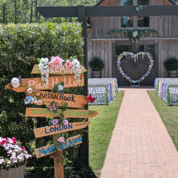 Wedding venue with a brick aisle, decorated with floral arrangements, leading to an outdoor ceremony area. A rustic wooden building with a heart-shaped floral wreath decorates the door, and a signpost with multiple colorful directional signs is in the foreground.