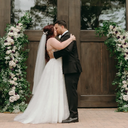 Bride and groom embracing in front of a wooden door decorated with white and purple flowers.