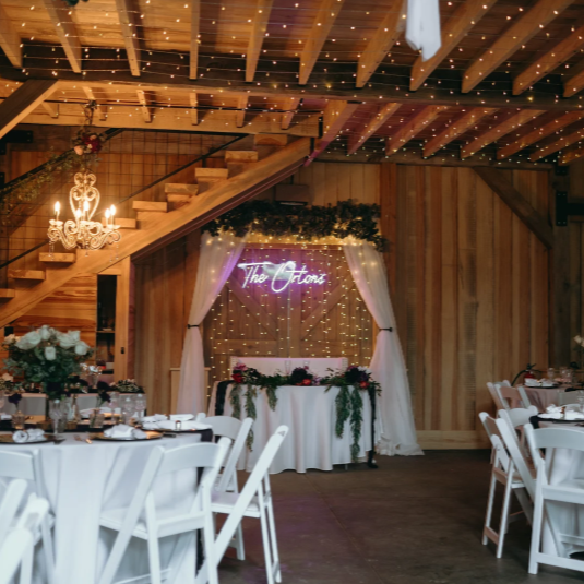 Wedding reception area with decorated tables, floral arrangements, and a neon sign that says 'The Ozans' in a rustic barn-style venue with wooden walls, string lights, and a staircase.
