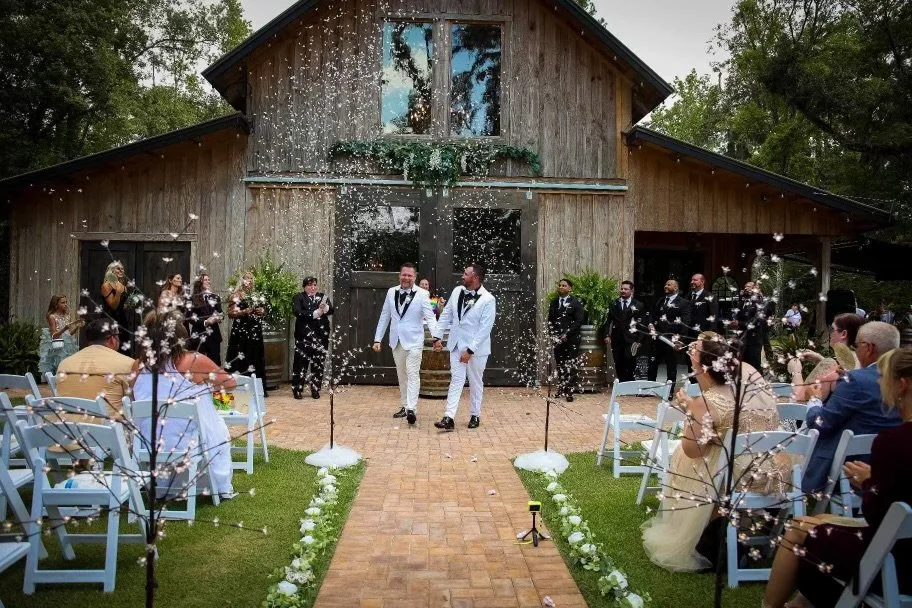 A wedding celebration outside a rustic barn with two grooms in white tuxedos walking down the aisle, surrounded by guests seated on white chairs and confetti in the air.