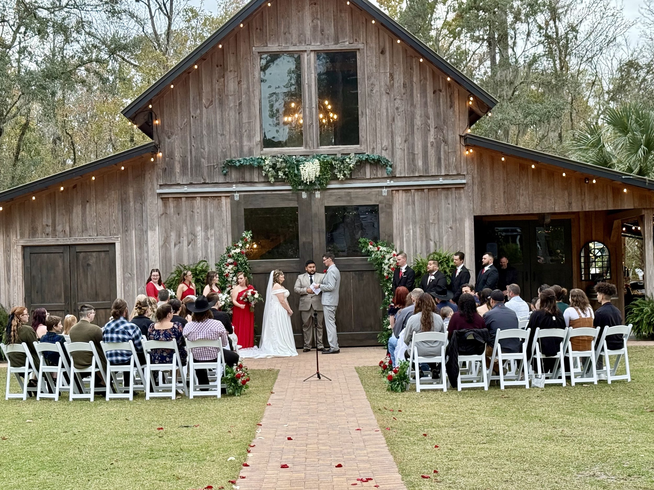 Outdoor wedding ceremony in front of a rustic wooden barn with string lights, floral decorations, and guests seated on white chairs.