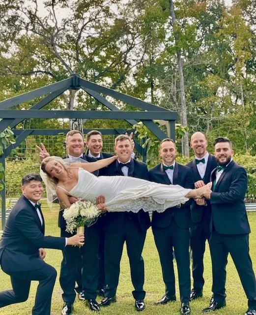 Groom lying on a group of seven groomsmen outdoors at a wedding, holding a bouquet and making peace signs.