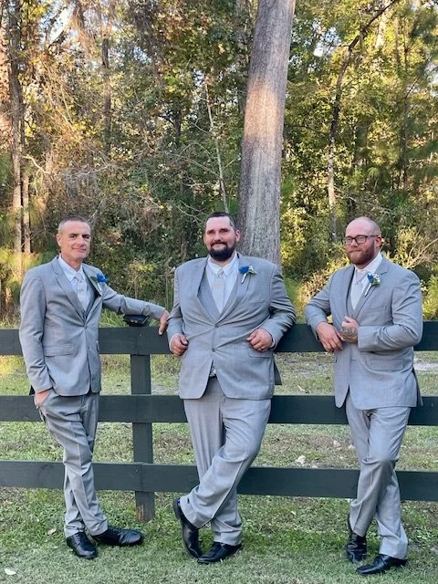 Three men in gray suits with blue boutonnieres posing outdoors in front of a wooden fence and trees.