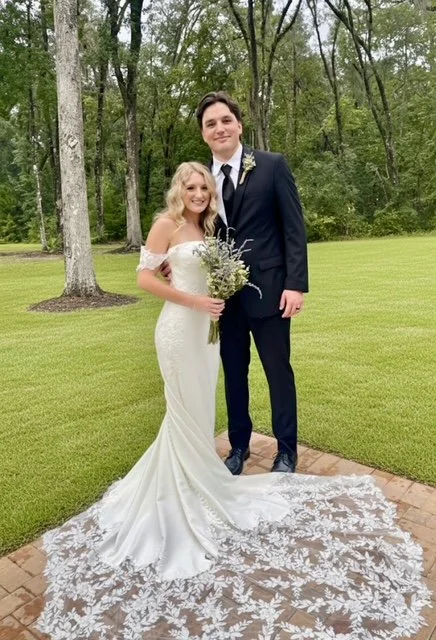 Bride and groom standing outdoors on a grassy lawn, with the bride holding a bouquet of flowers and wearing a white wedding gown with lace train, and the groom in a black suit and tie, both smiling.