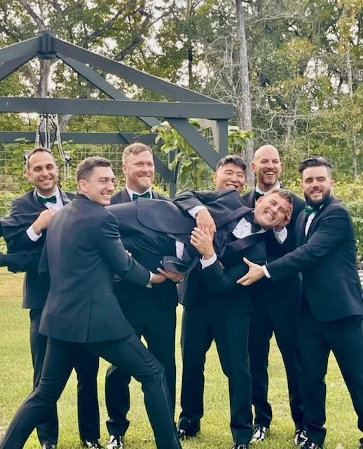 Group of seven men in tuxedos smiling and lifting another man in a tuxedo outdoors in a garden or backyard setting.