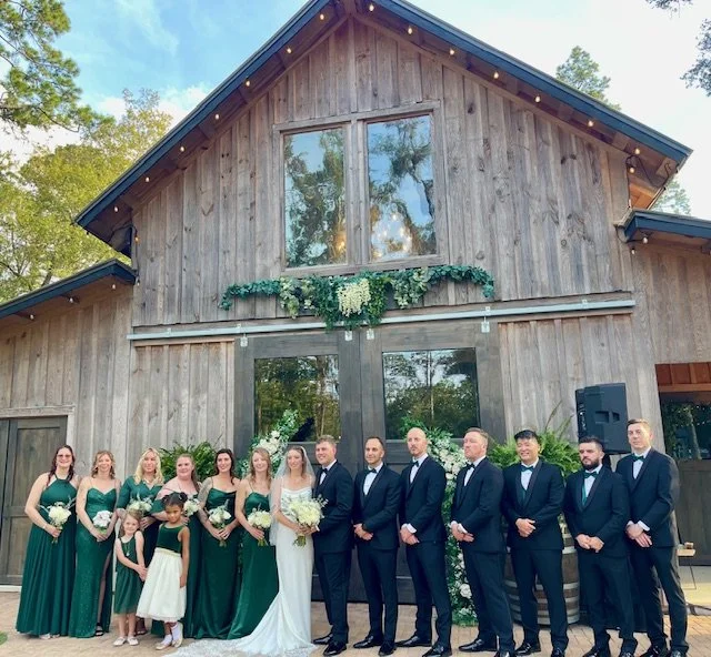 Group of people dressed in formal attire on wedding day in front of wooden building with greenery and string lights