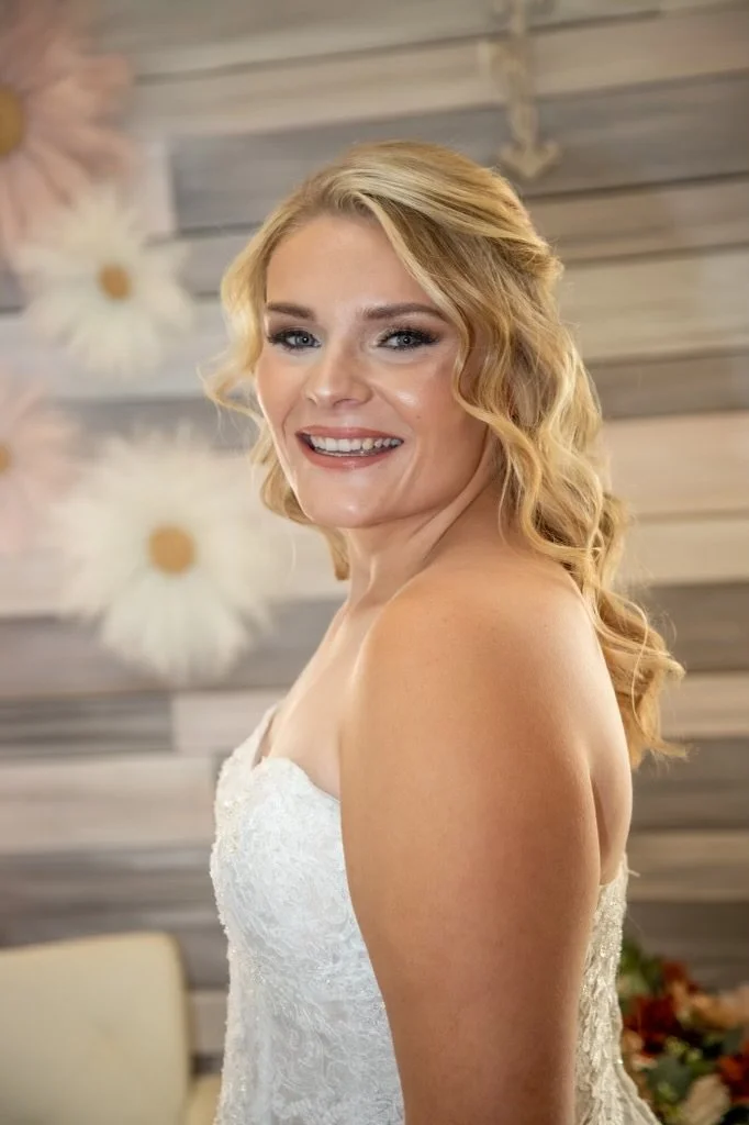 A blonde woman with wavy hair smiling at a wedding or formal event, wearing a white lace strapless dress with a wooden wall and decorative flowers in the background.
