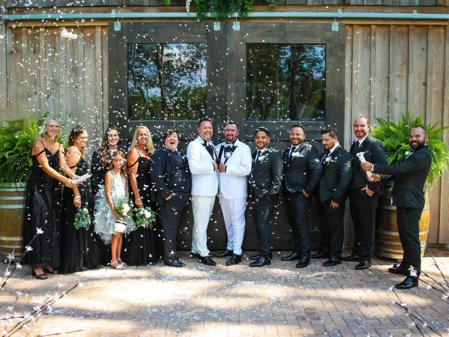 A wedding celebration with a diverse group of people, including the bride and groom in white tuxedos, surrounded by friends and family, with confetti falling and a wooden building in the background.