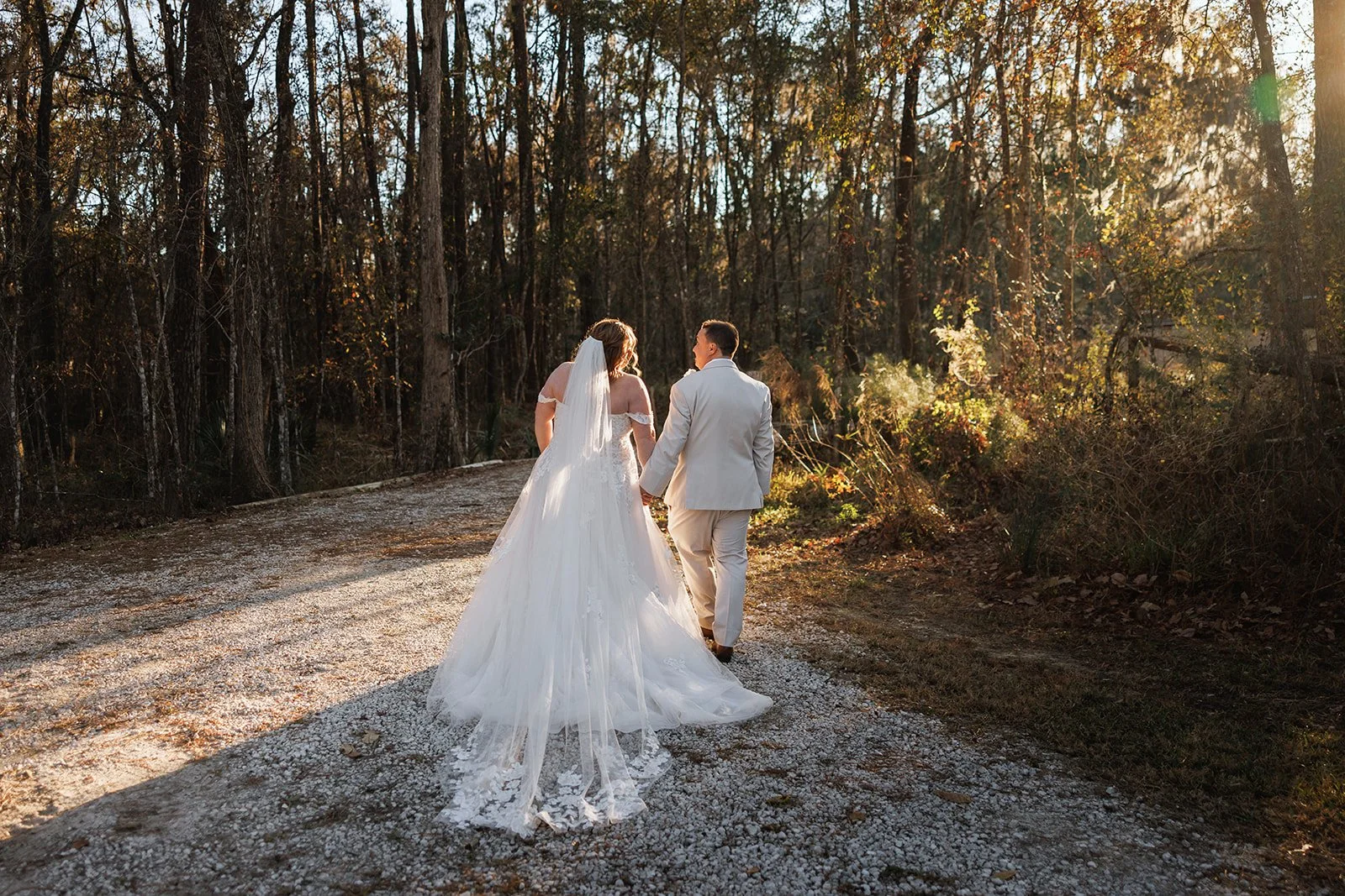 A bride and groom walking hand in hand on a dirt path in a wooded area during sunset, with the bride wearing a white wedding gown and veil, and the groom in a light-colored suit.
