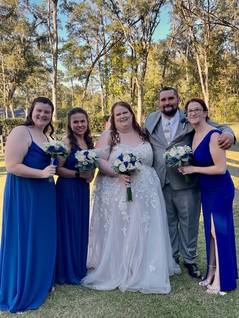 A group of five people at a wedding outdoors in a wooded area, including a bride in a white wedding gown, a groom in a gray suit, and three bridesmaids in blue dresses, all holding bouquets of flowers.