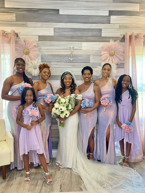Seven women, including bride in white gown holding bouquet, and six bridesmaids in matching lavender dresses, pose for a photo in a decorated indoor setting with pink flowers and pastel curtains.