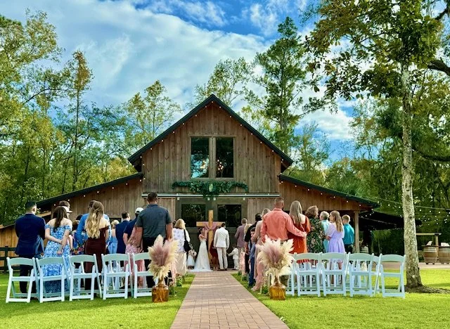 People attending a wedding ceremony outside a rustic wooden barn, with chairs arranged on either side of a brick aisle, in a green outdoor setting with trees and a partly cloudy sky.