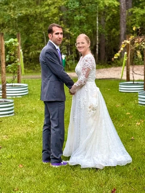 A bride and groom holding hands and standing outdoors in a grassy area with trees and greenery in the background, during their wedding.