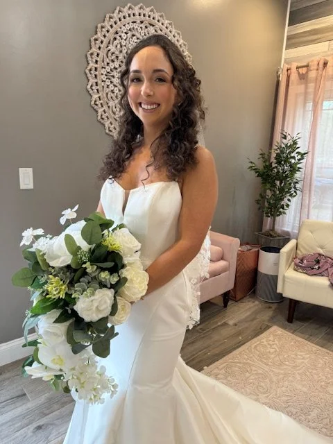 A woman in a white wedding dress holding a large bouquet of white and green flowers indoors.