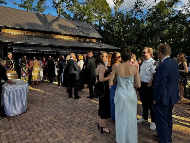 People dressed in formal attire gather outdoors near a rustic building with a metal roof, socializing at an event on a brick-paved area.